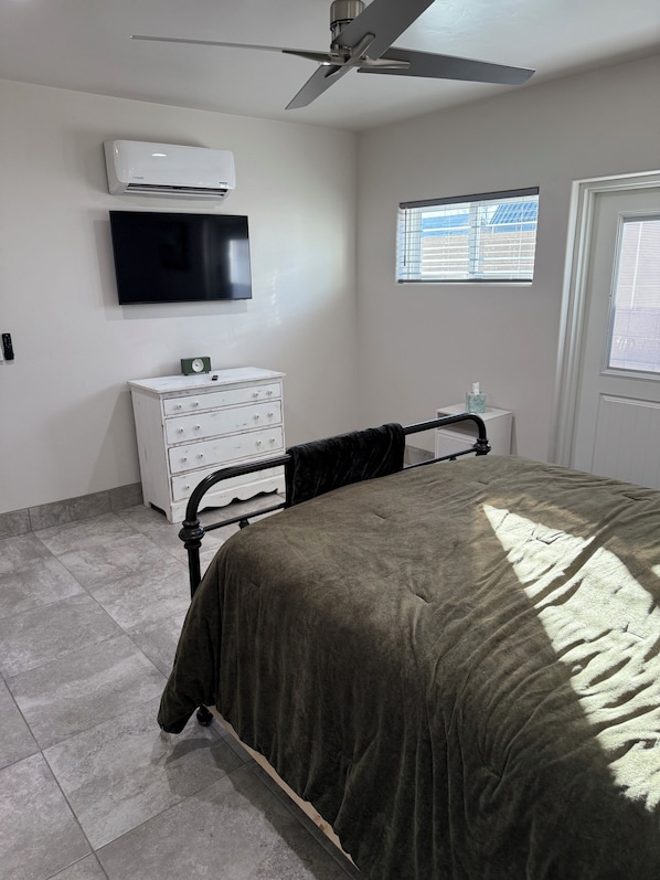 A bedroom with a bed, a white dresser, a television, an air conditioner, a ceiling fan, and a window with blinds.