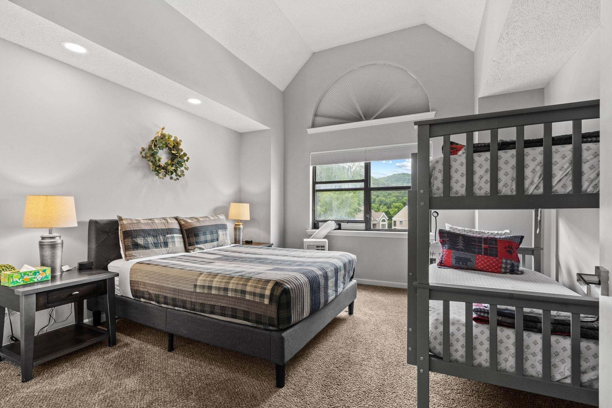 A bedroom with a bunk bed, a bed with a plaid comforter, a desk with a lamp, a window with a view of greenery, and a wall-mounted wreath.