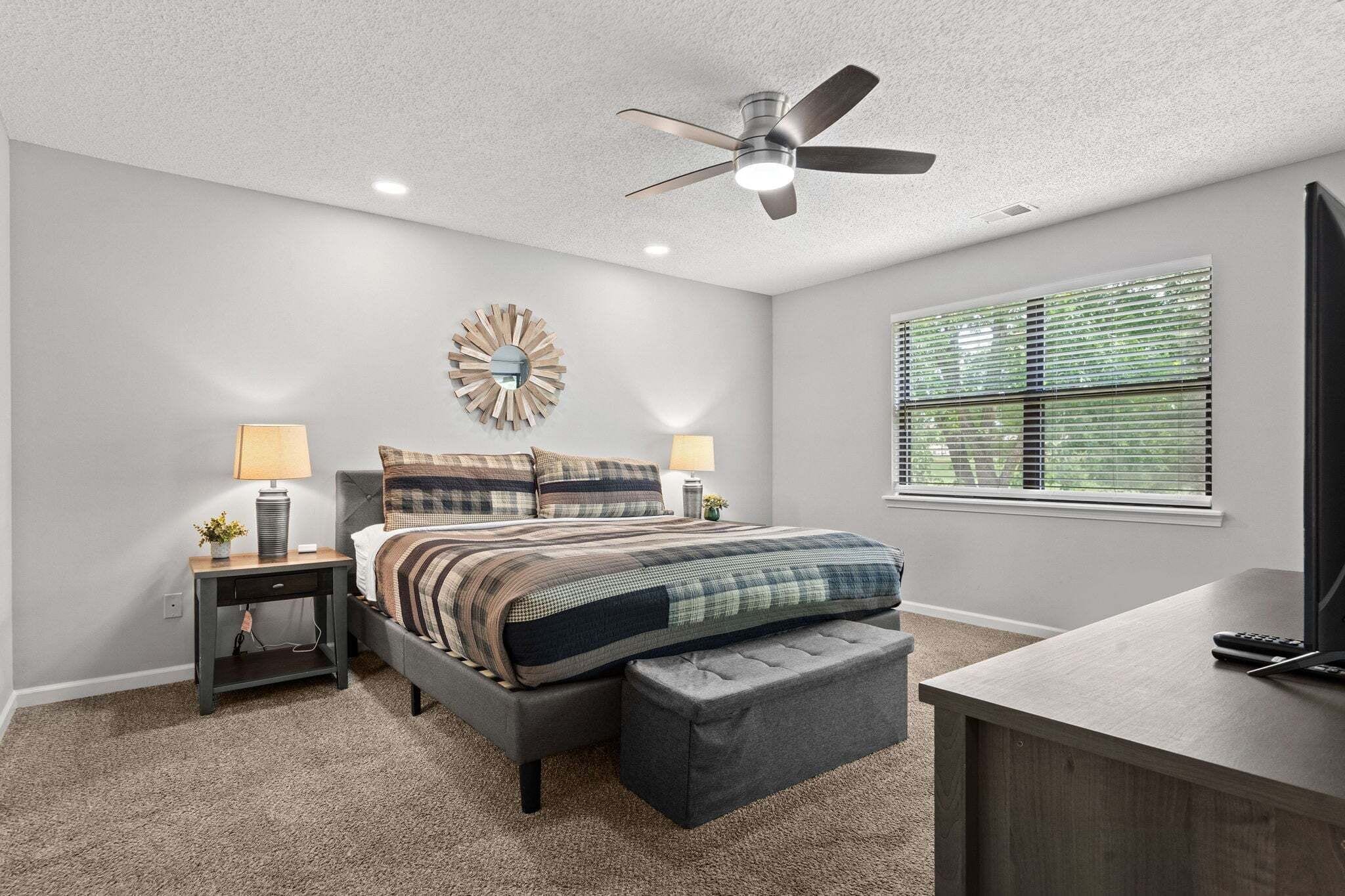 A bedroom with a bed, nightstands, a ceiling fan, a window with blinds, and a TV stand.