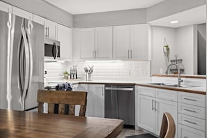 A modern kitchen with white cabinetry, stainless steel appliances, and a wooden dining table. - Caddy Shack | Pointe Royale 3BR Condo w/ Pools + Golf Views (PR 61-8) (Branson)
