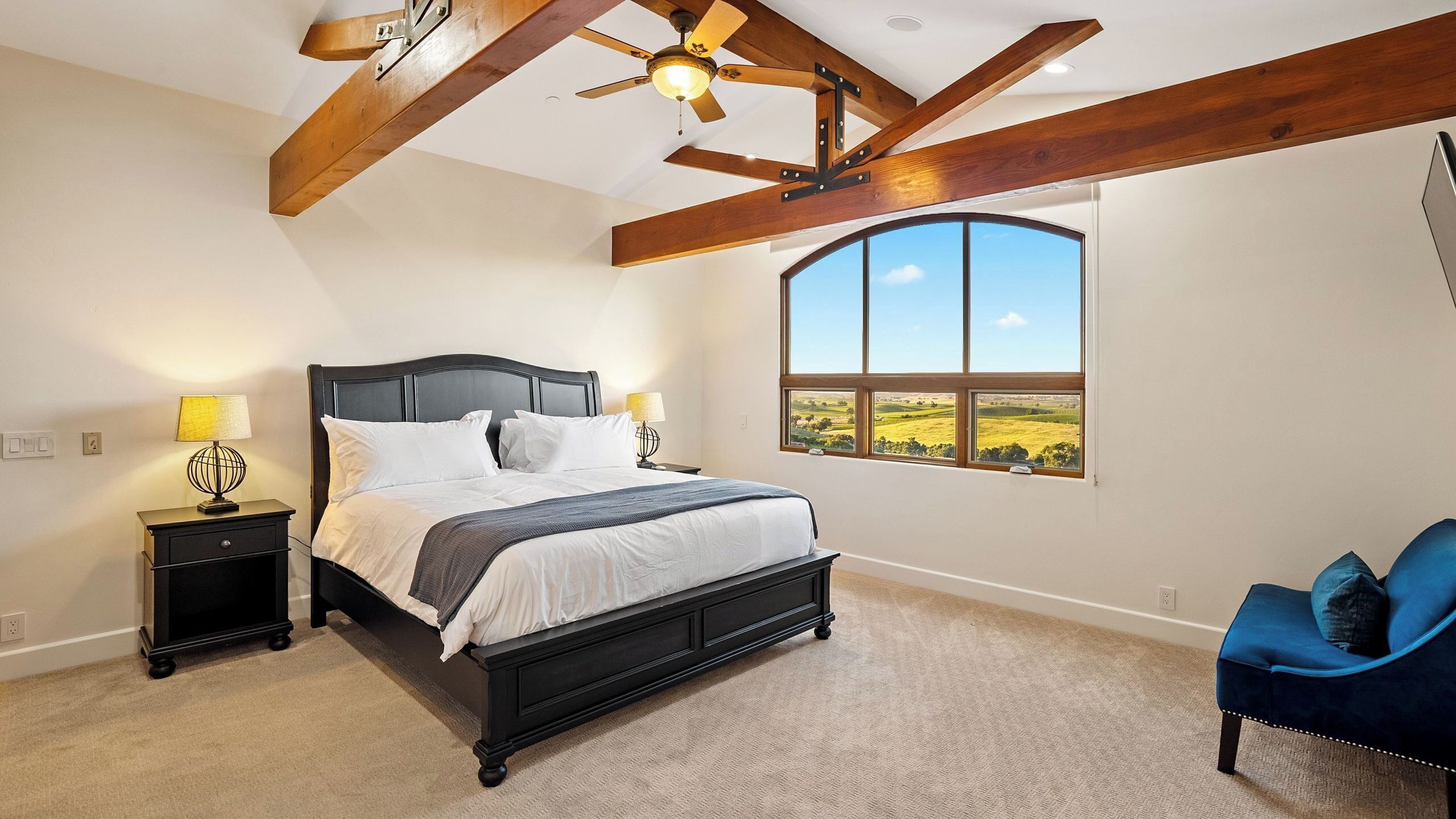 A bedroom with a large bed, two bedside tables, a ceiling fan, a large window with a view, and a blue chair.