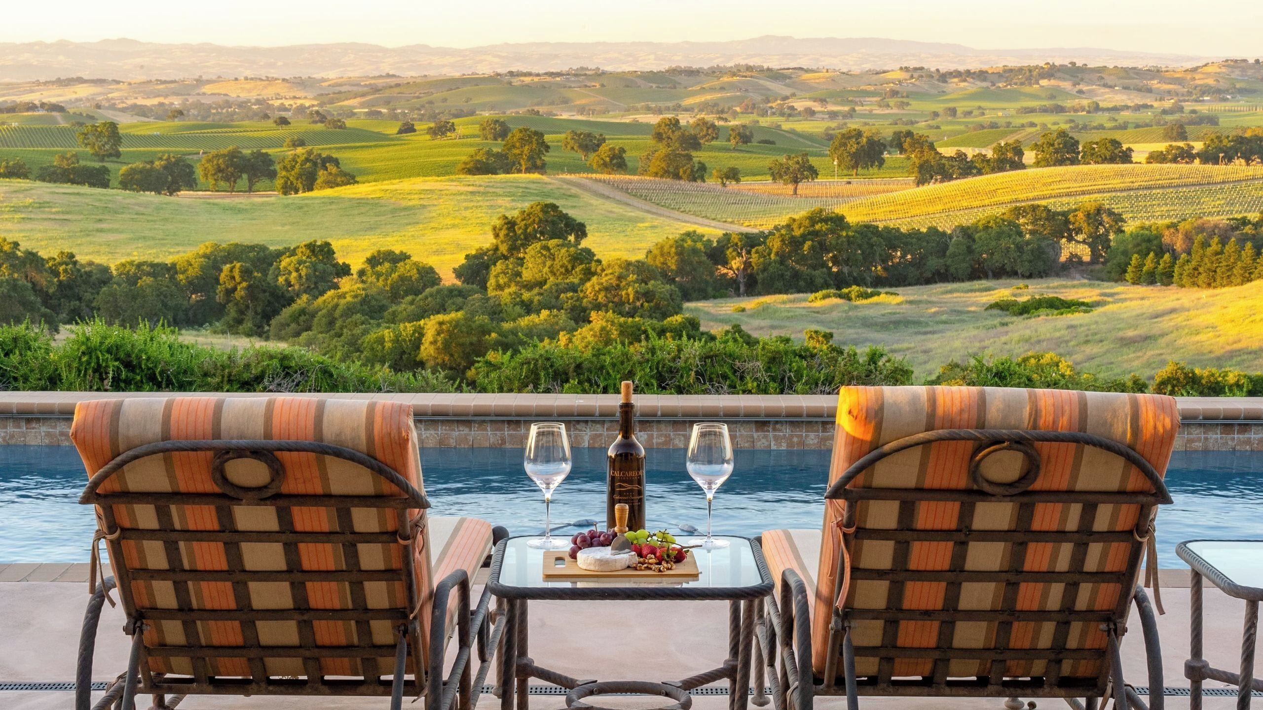 A poolside setting with two chairs, a table with a bottle of wine, glasses, and a fruit platter.