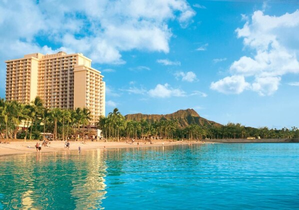Een hotel aan het strand met een zwembad, palmbomen en een berg op de achtergrond.
