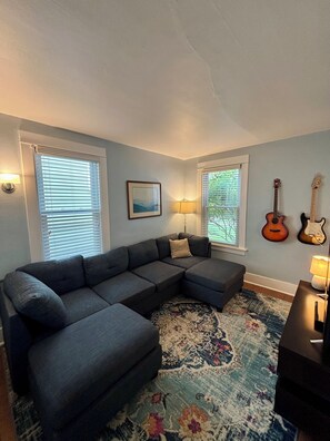 Ein Wohnzimmer mit einem Ecksofa, zwei an der Wand befestigten Gitarren und einem gerahmten Bild.