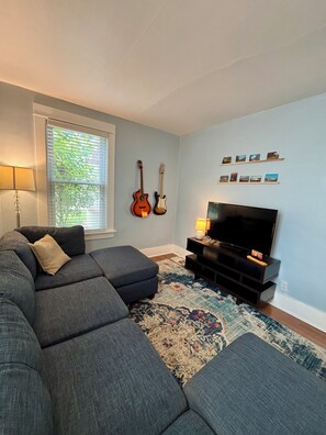 Una sala de estar con un sofá modular, un televisor de pantalla plana, una guitarra y una ventana con persianas.
