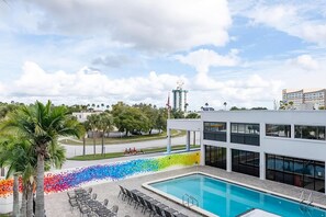 Ein Schwimmbecken mit Liegestühlen, eine farbenfrohe Mosaikwand und Blick auf eine Stadtlandschaft mit Gebäuden und einem hohen Bauwerk.