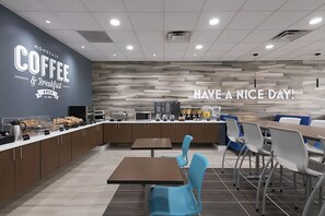 Ein modernes Café mit einer Theke, die Gebäck und Kaffeemaschinen präsentiert, Tischen und Stühlen zum Sitzen sowie einer Wand mit den Aufschriften "MONDELE COFFEE & BREAKFAST AREA FL USA" und "HAVE A NICE DAY!".