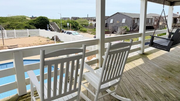 Une terrasse en bois avec des fauteuils à bascule blancs, donnant sur une piscine et une zone sablonneuse.