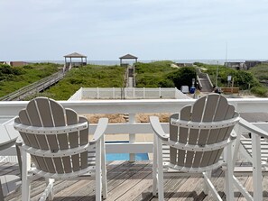 Eine Terrasse mit zwei Adirondack-Stühlen, die auf einen Strand, einen Pool und einen weißen Zaun, der zu einem Promenadenweg führt, ausgerichtet ist.