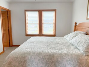 A bedroom with a bed, a window with blinds, a wooden door, and a headboard. - The Dune Abides - Oceanfront, Private Yard with Pool, Hot Tub, Volleyball Court (Nags Head)