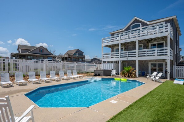 A swimming pool with lounge chairs and a two-story building with a balcony. - The Dune Abides - Oceanfront, Private Yard with Pool, Hot Tub, Volleyball Court (Nags Head)
