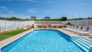 Une piscine rectangulaire à l’eau d’un bleu limpide, entourée d’une clôture blanche. On aperçoit des chaises longues et un kiosque au fond.