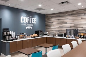 Ein moderner Bereich für Kaffee und Frühstück mit einer Theke, auf der Kaffeemaschinen, Gebäck und eine Kaffeestation präsentiert werden. Es gibt Tische und Stühle zum Sitzen.