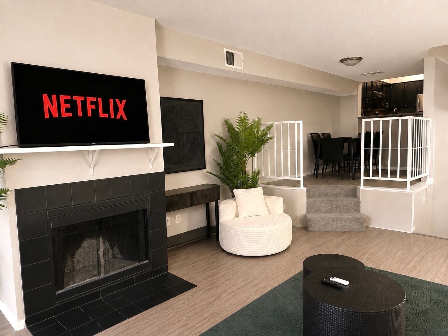 A modern living room with a fireplace, a flat-screen TV mounted on the wall, a white armchair, a small round table, and a green rug.