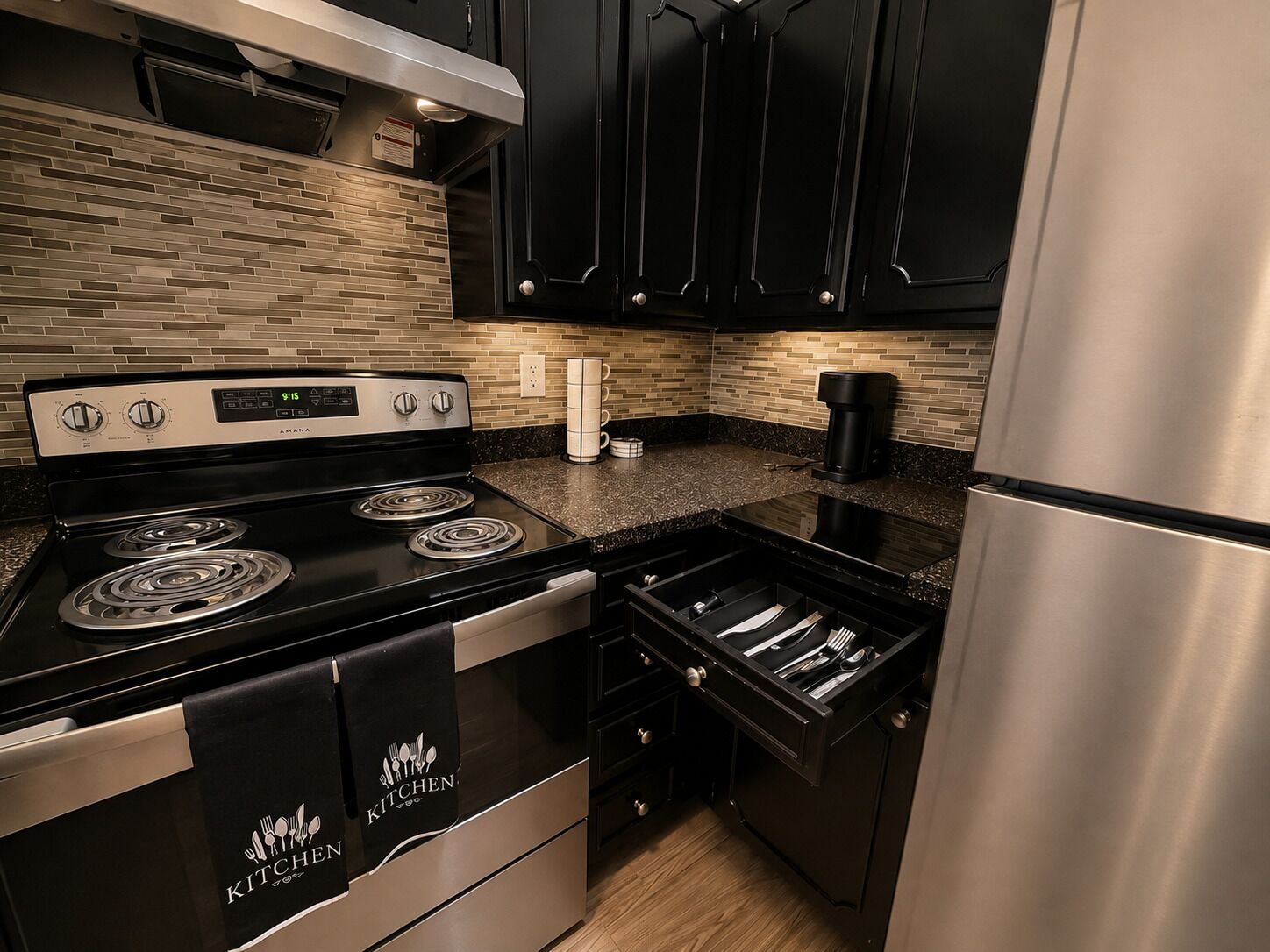 A modern kitchen with black cabinetry, a stainless steel refrigerator, a black stove, and a coffee maker.