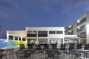 Eine Außenpatio-Terrasse mit Tischen und Stühlen, ein Schwimmbad und ein Mehrstöckiger Gebäudebau mit Balkonen.
