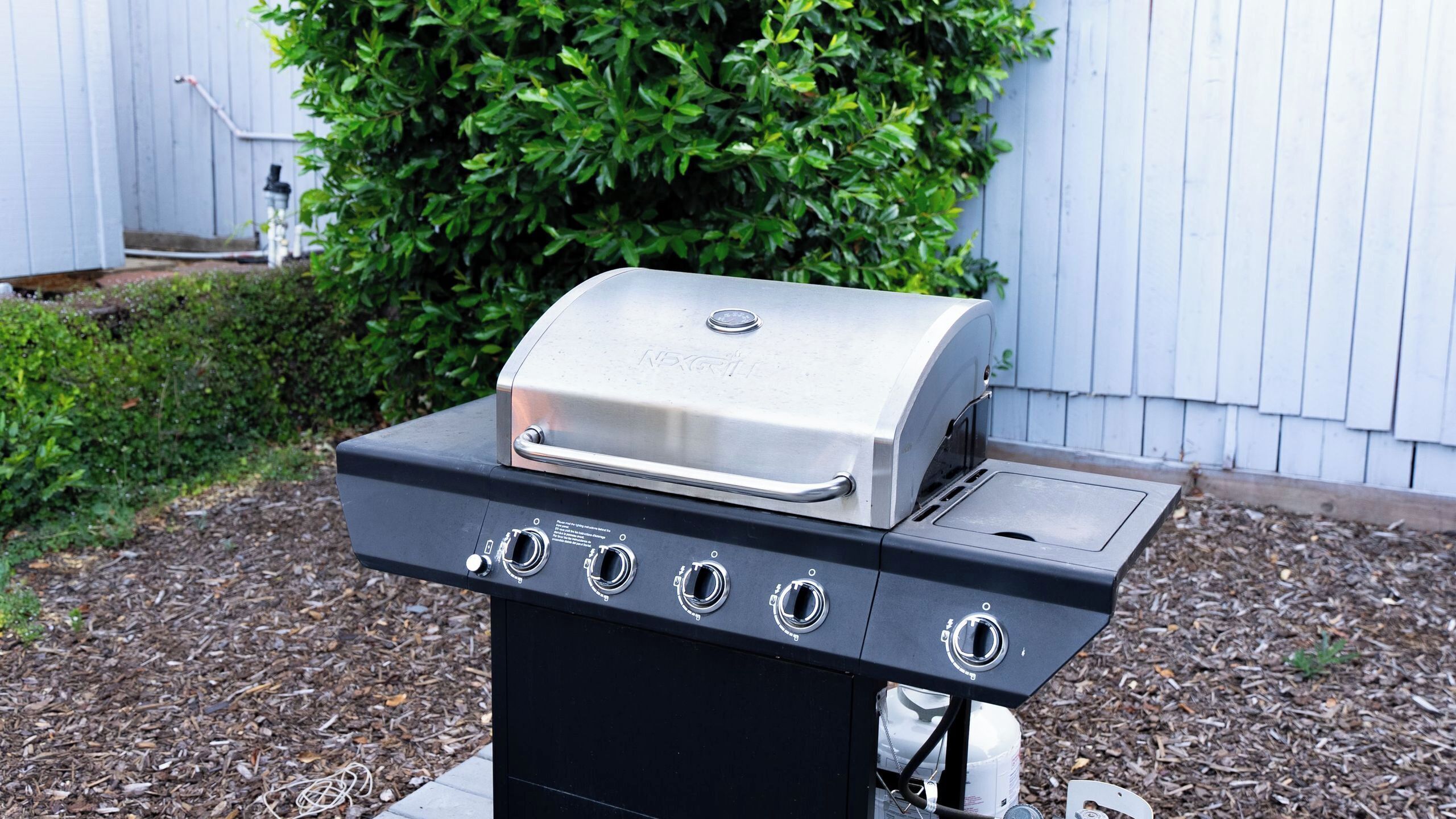 A gas grill with four burners and a side shelf, set on a wooden deck with mulch.