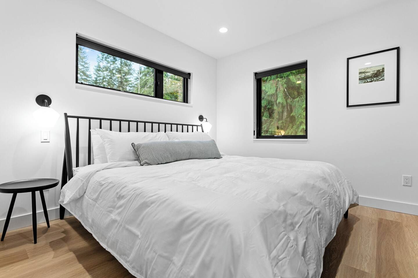 Une chambre à coucher avec un lit, deux fenêtres, une table de chevet et un tableau encadré au mur.