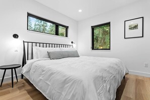 Un dormitorio con cama, dos ventanas, una mesita de noche y un cuadro en la pared.