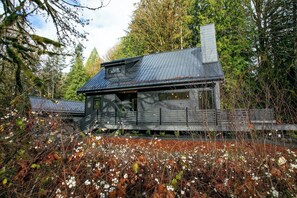 Una cabaña con techo de metal, rodeada de árboles y un jardín con flores blancas.
