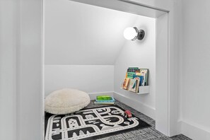 A small, white-walled room with a white fluffy chair, a black and white patterned rug, a toy car, and a wall-mounted book rack with books. - Fern + Cedar Cabin | hot tub | fireplace | firepit (Deming)