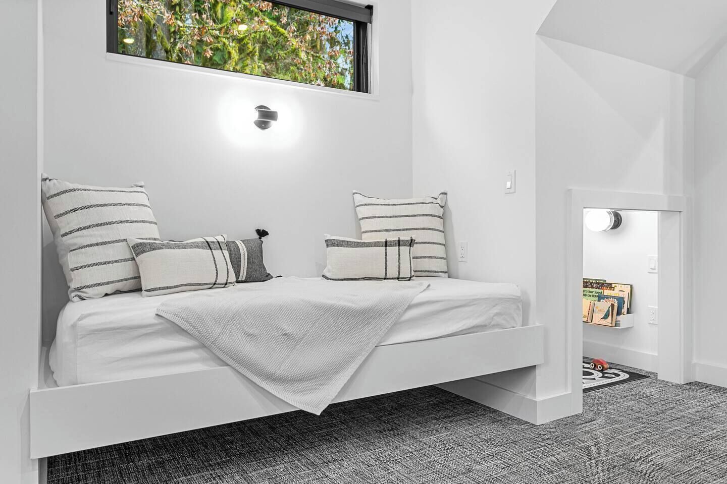 A white room with a bed, striped pillows, a window with a view of greenery, and a door leading to another room.