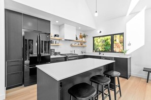 A modern kitchen with a central island, bar stools, and a large window with a view of greenery. - Fern + Cedar Cabin | hot tub | fireplace | firepit (Deming)