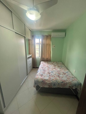 Ein kleines Schlafzimmer mit einem Bett, einem Fenster mit Vorhängen, einem Deckenventilator und einer Klimaanlage.