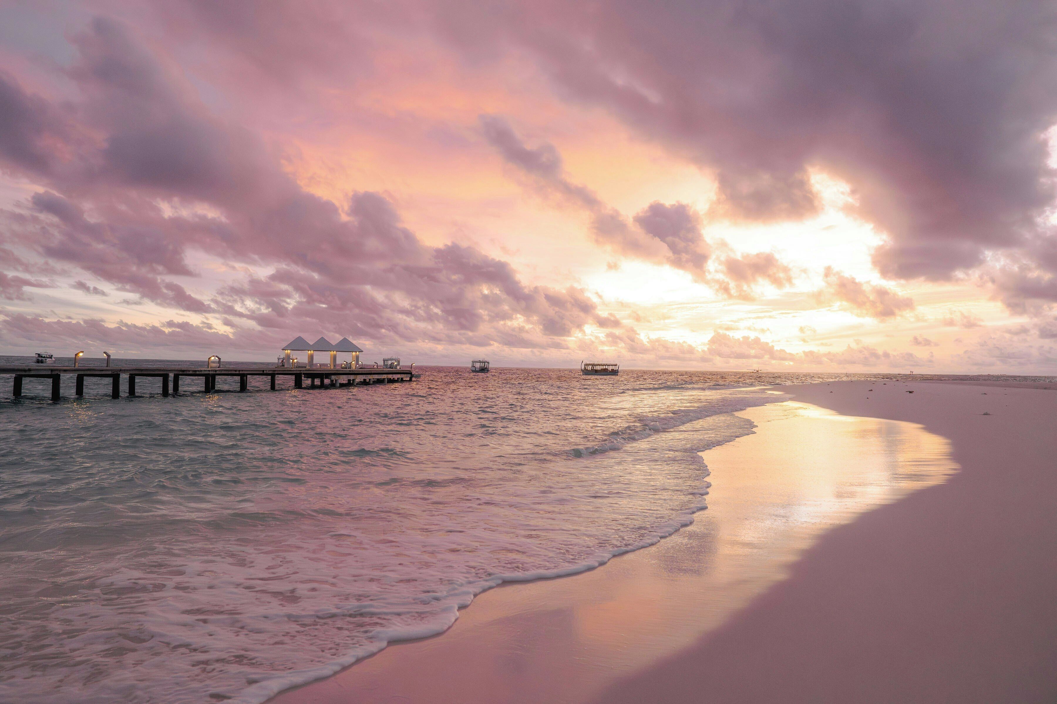 Una playa con un muelle que se adentra en el agua, un barco y una puesta de sol.