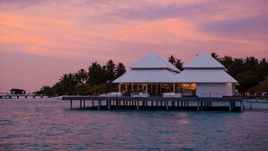 Et resort med to hvide overvandsbungalows forbundet af en dok, omgivet af palmer og et roligt hav.