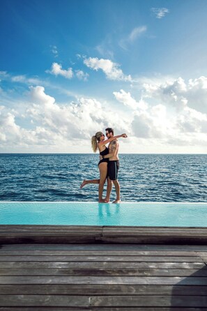 Una pareja se abraza en una terraza de madera con vista a una piscina y al océano.