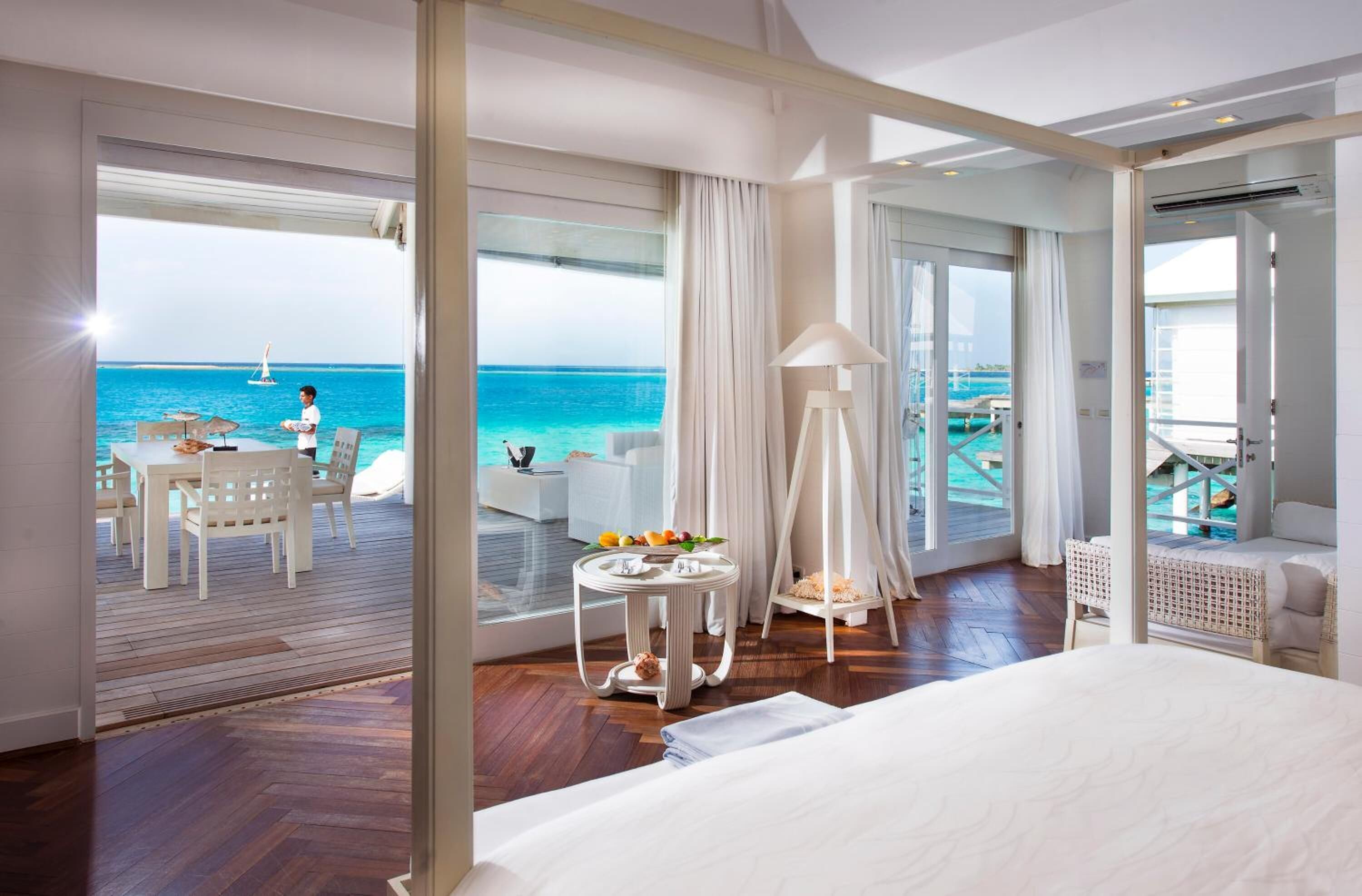 Habitación de hotel con una cama grande, una zona de comedor con mesa y sillas, una mesita redonda con lámpara y vista al océano.