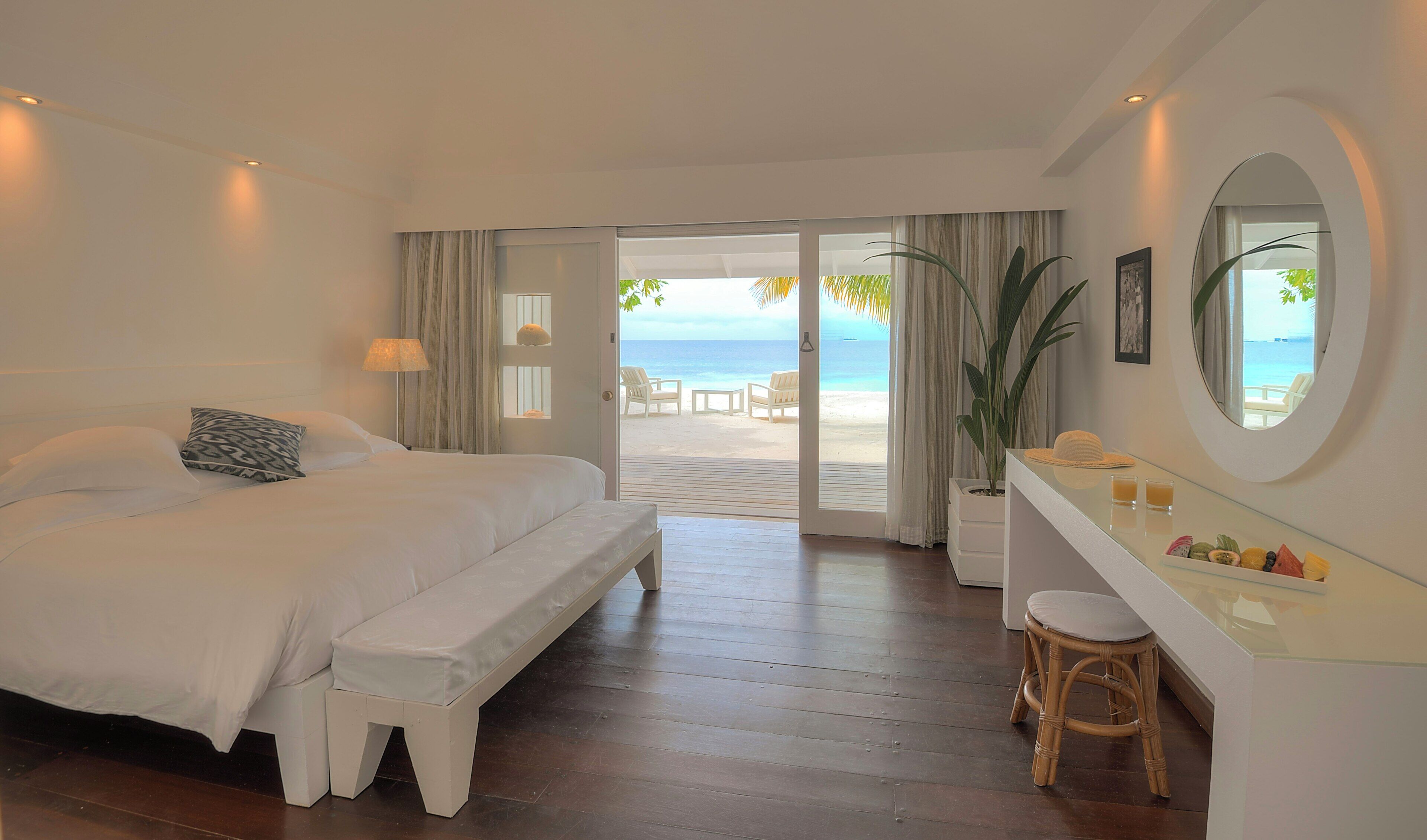 Habitación de hotel con cama, banco, mesita de noche y vista al mar.