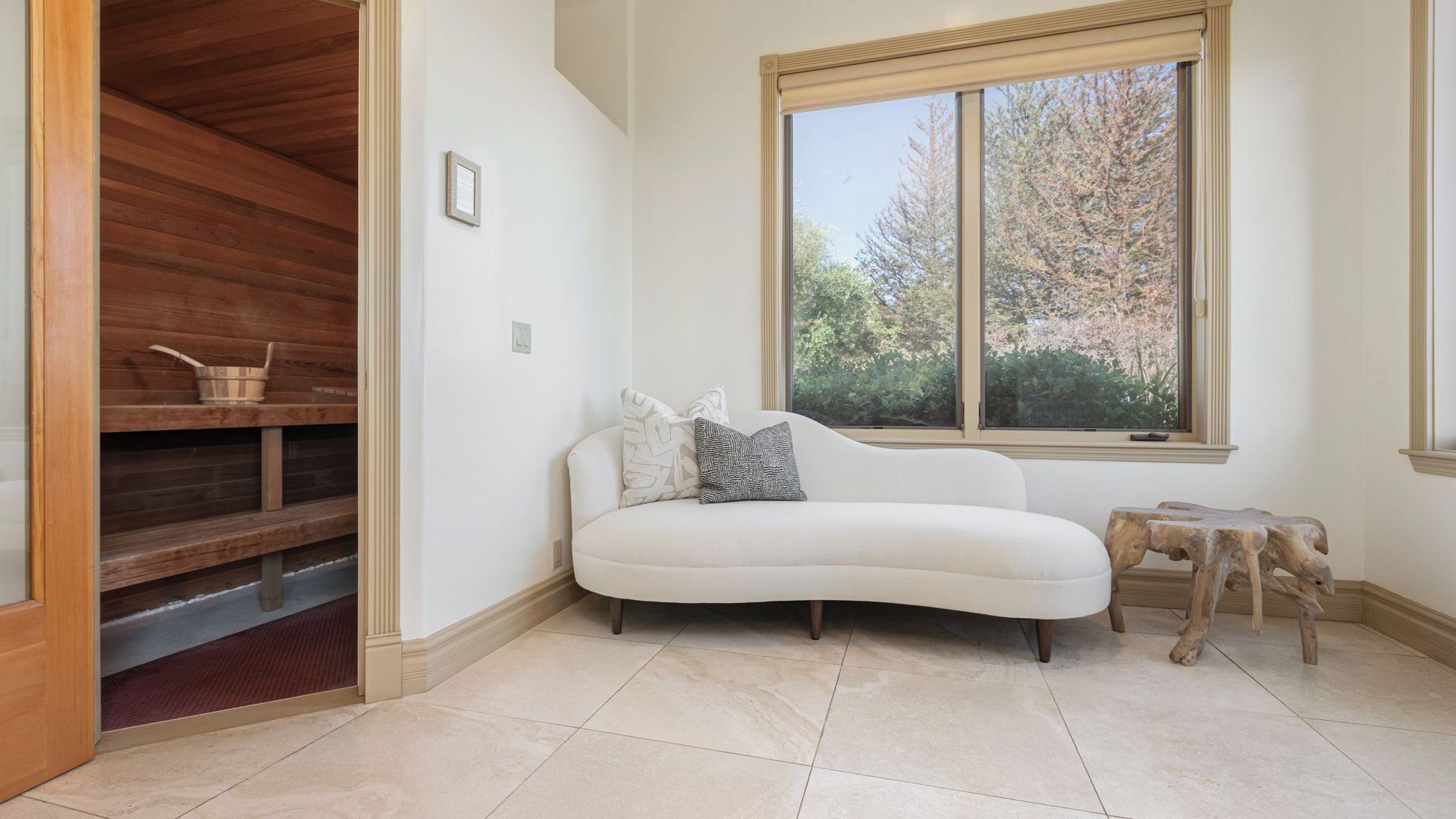 A room with a white sofa, a wooden bench, a window with a view of trees, and a tiled floor.
