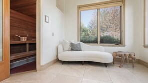 Ein Zimmer mit einem weißen Sofa, einer hölzernen Bank, einem Fenster mit Blick auf Bäume und einem Fliesenboden.