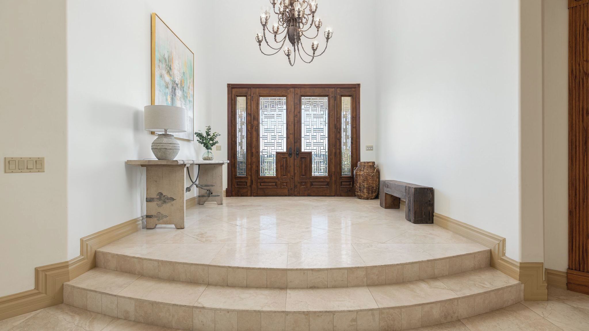 A spacious foyer with a marble staircase, a chandelier, a wooden door with glass panels, and a framed artwork on the wall.