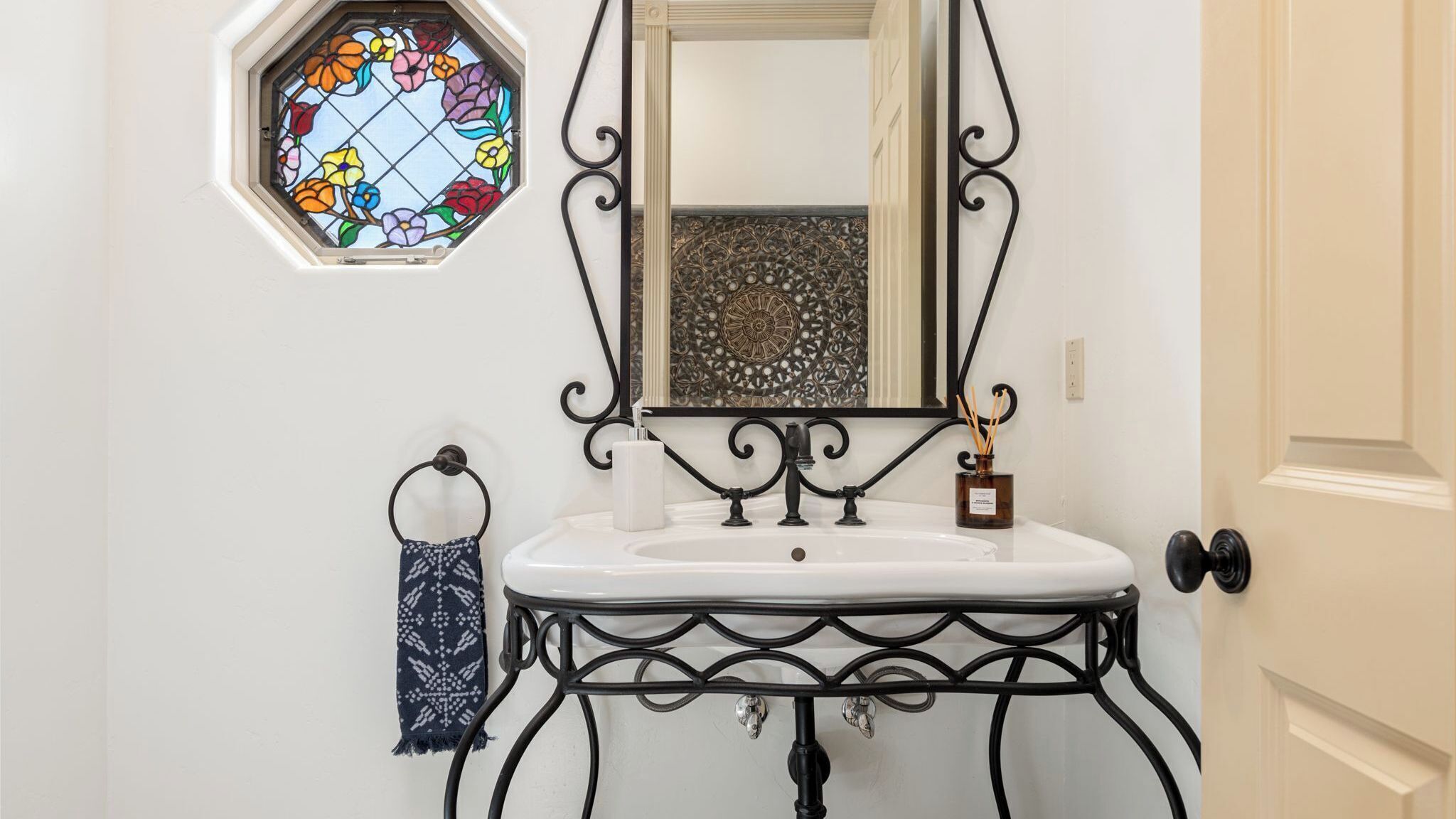 A bathroom with a white sink, black iron frame, a stained glass window, and a mirror above the sink.