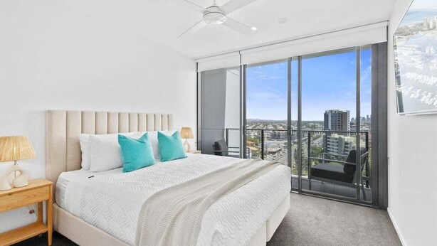 A modern bedroom with a large bed, a ceiling fan, and a city view through floor-to-ceiling windows. - Sandbar Burleigh - Ocean Delight (Gold Coast)
