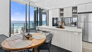 A modern kitchen with a view of the ocean, a dining area with a round table and chairs, and a balcony with a view. - Sandbar Burleigh - Ocean Delight (Gold Coast)