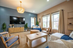 Ein Wohnzimmer mit Flachbildfernseher, Holzmöbeln, einem gewebten Hocker und großen Fenstern.