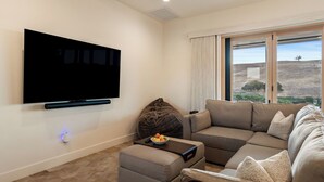 Ein Wohnzimmer mit flachbildfernseher an der Wand, einem Ecksofa, einem Sitzsack und einem kleinen Tisch mit Obstschale.