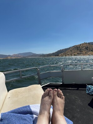 A boat deck with a person's feet resting on a towel, water in the background, and a hilly landscape.