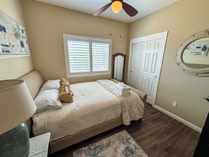 Une chambre à coucher comprenant un lit, un ventilateur de plafond, une fenêtre avec des stores, une porte, un miroir et un tapis.