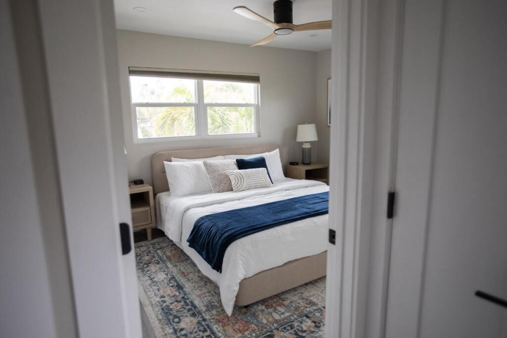 A bedroom with a bed, nightstands, a lamp, a ceiling fan, and a window with a view of greenery.