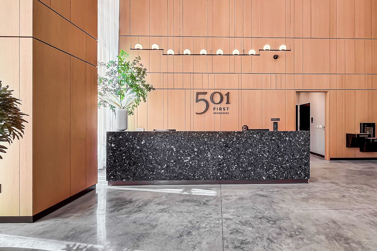 A reception desk with a black marble top, a wooden wall with the number '501' and 'First Residences' sign, and a potted plant on the left.