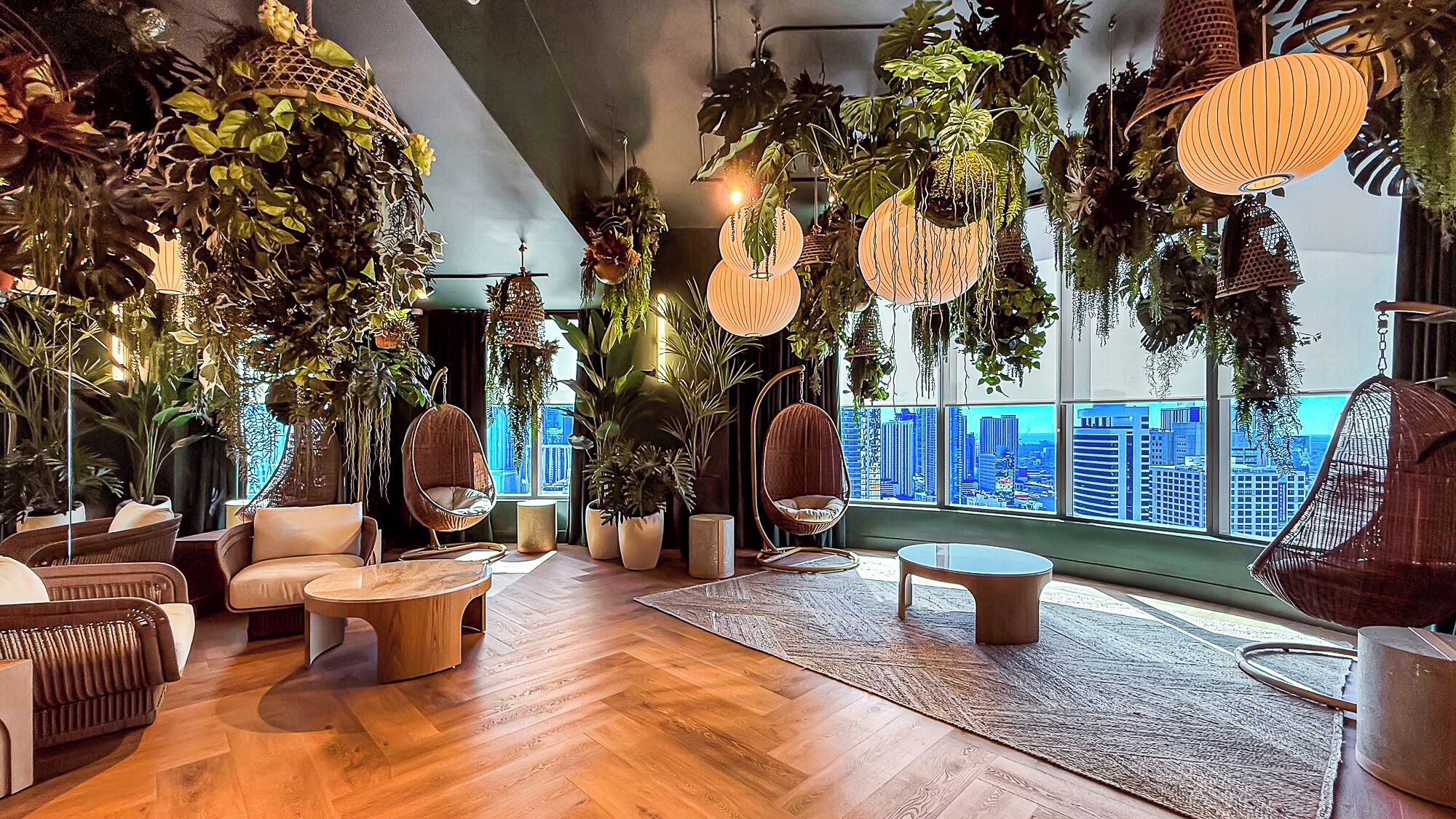 A modern lounge with hanging plants, wicker furniture, and a city view.