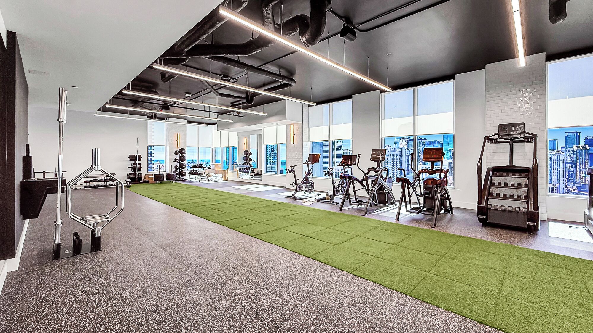 Un gimnasio con equipo de ejercicio, una zona de césped artificial y amplios ventanales con vistas a la ciudad.