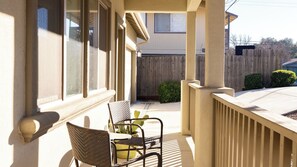 Eine überdachte Veranda mit zwei Korbstühlen und einem Blumentopf, ein Holzzaun im Hintergrund und ein Gebäude mit Fenster und Tür.