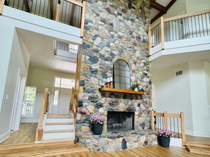 A spacious interior with a stone fireplace, wooden staircase, and a balcony with a railing.