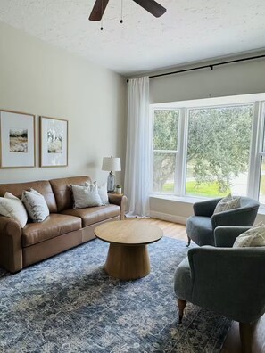 A living room with a brown leather sofa, a round wooden coffee table, and a grey armchair. There are two framed pictures on the wall, a lamp on a side table, and a large window with curtains.